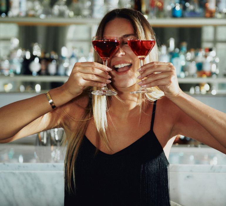 A woman is holding two cocktails infront of her face 