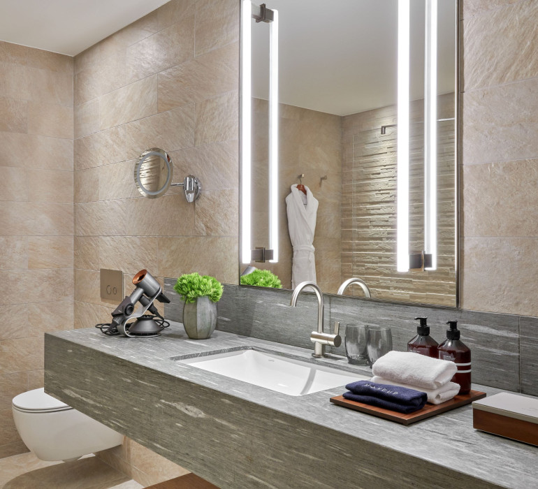 Bright bathroom with a stone sink and beige tiles in front of a light-up mirror