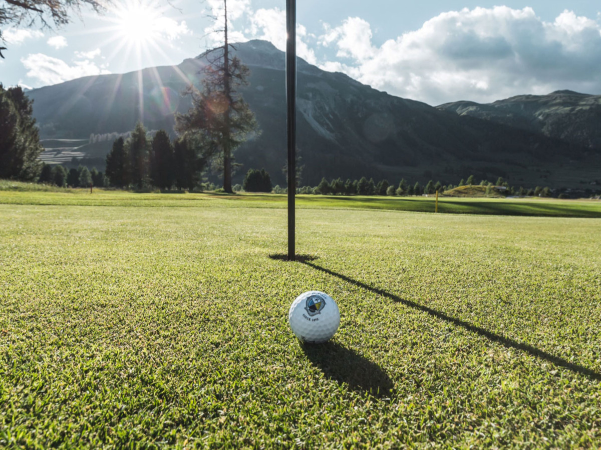 A golf ball on green grass with mountains and the sun in the background